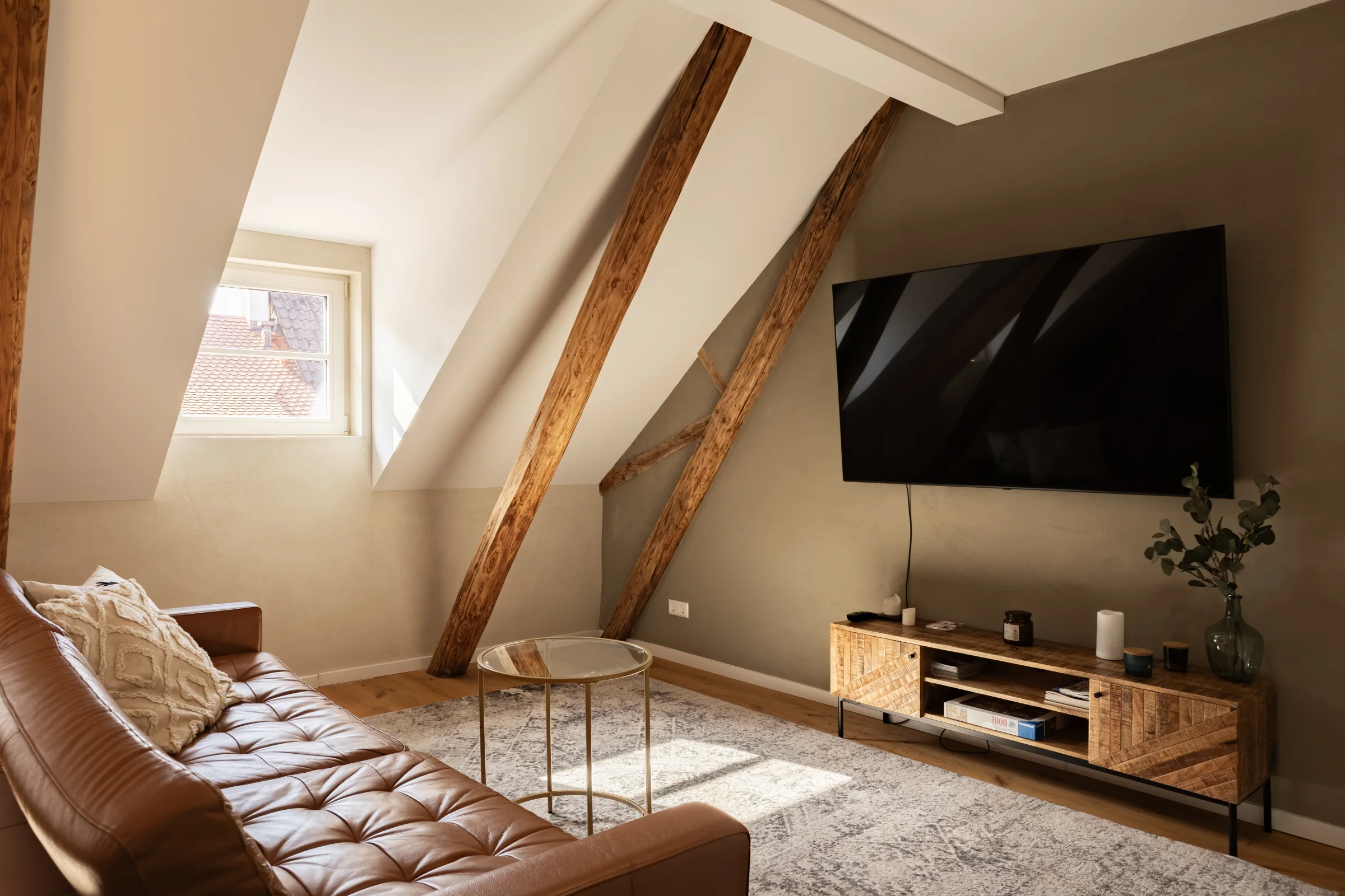 Sitzecke unter Dachschräge mit sichtbaren Holzbalken, braunem Sofa und wandmontiertem Fernseher